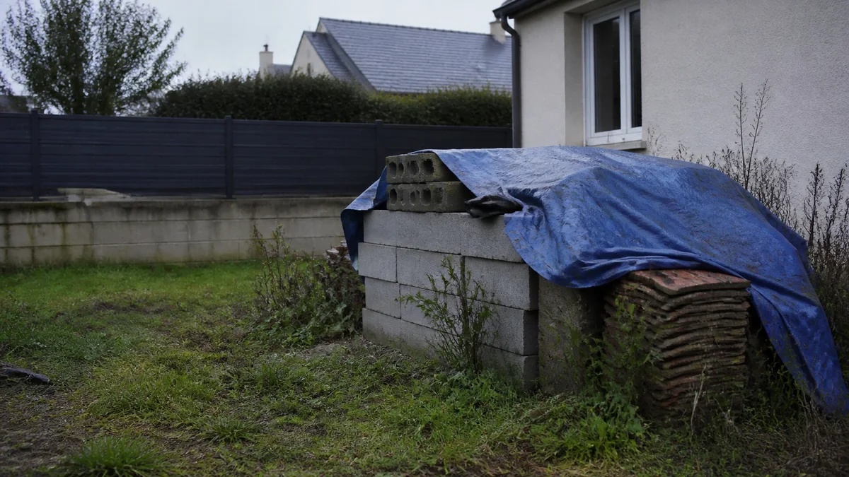 Pile de parpaings et de tuiles recouverte d'une bâche bleue dans un jardin de pavillon francilien, chantier laissé à l'abandon