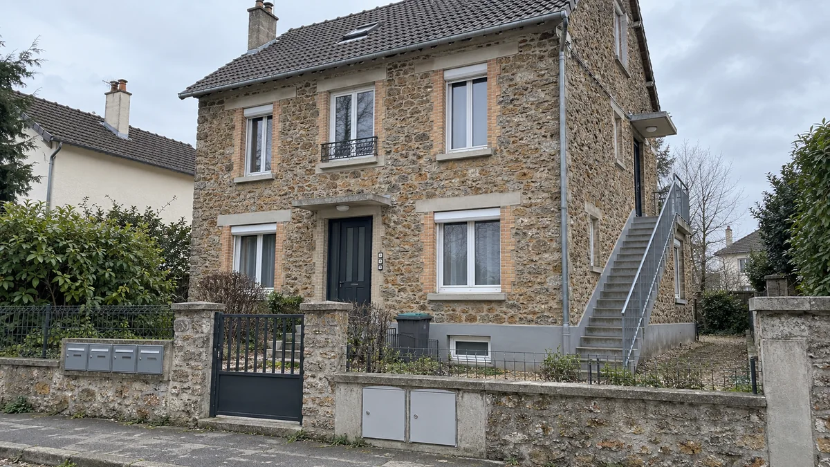 Pavillon en meulière de banlieue parisienne divisé en logements : quatre boîtes aux lettres sur le muret, escalier extérieur en acier ajouté sur le côté, fenêtre neuve au sous-sol