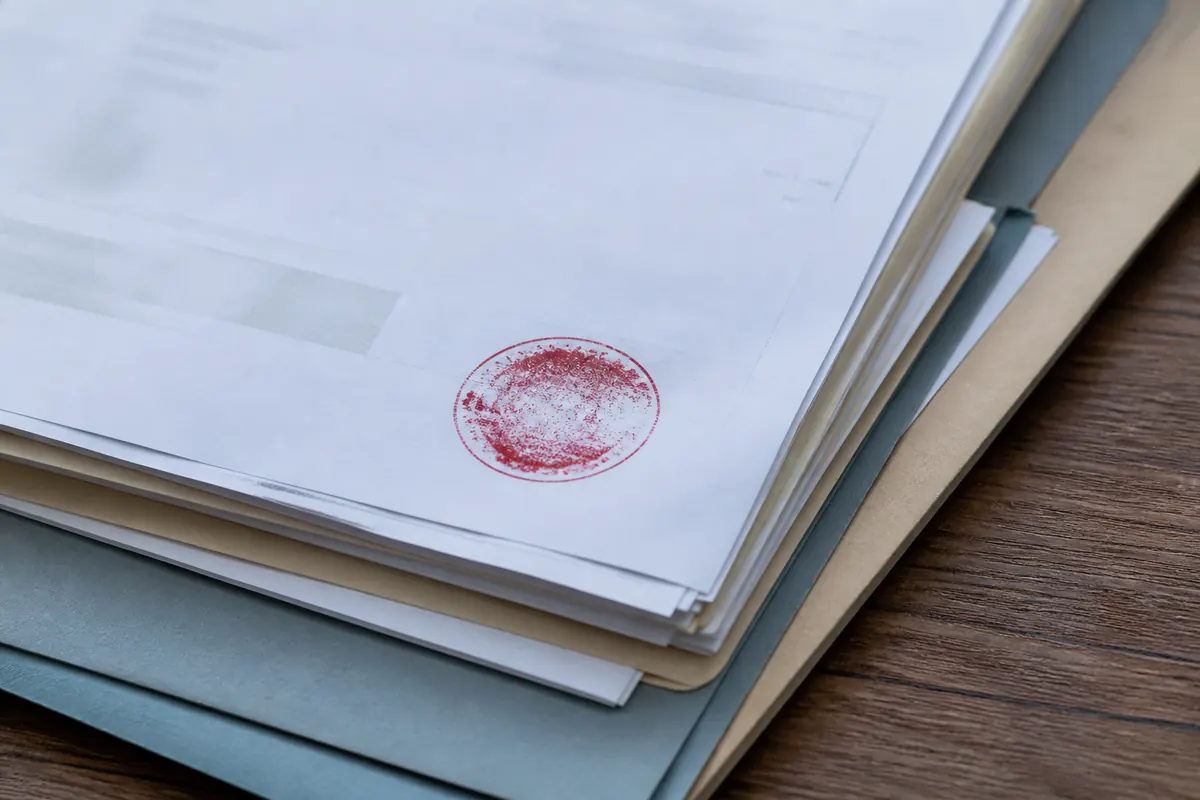Pile de documents et de dossiers de sinistre sur un bureau avec un tampon encreur circulaire flouté posé dessus, lumière froide naturelle