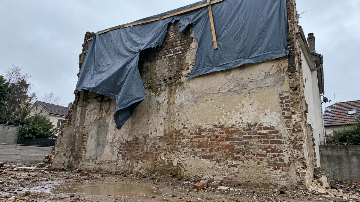 Haut d'un mur pignon mis à nu après une démolition : la bâche plastique gris-bleu fixée par des tasseaux est déchirée par le vent, un pan entier pend, un tasseau vertical tient de travers, la maçonnerie derrière la déchirure est sombre et mouillée, une flaque au pied du mur sur les gravats