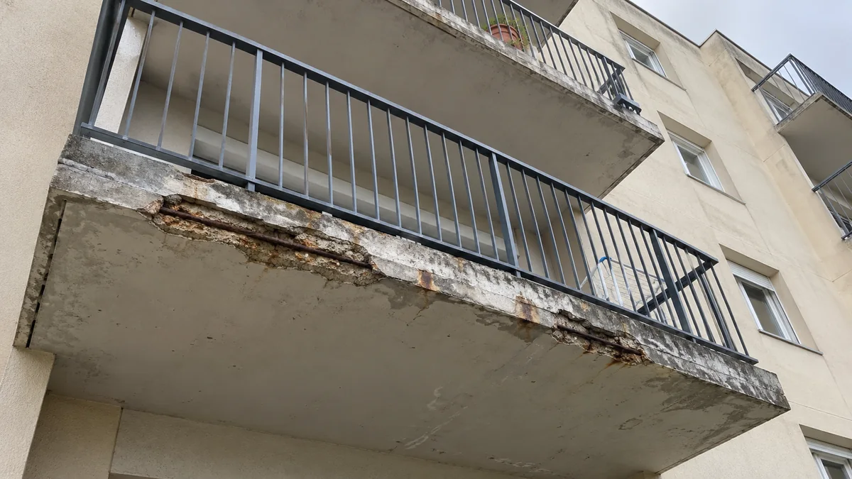 Dessous d'un balcon en béton d'une résidence des années soixante-dix vu d'en bas : le nez de dalle a éclaté sur plusieurs dizaines de centimètres et laisse voir des armatures rouillées, avec des coulures brunes sur la sous-face, sous un garde-corps métallique gris et d'autres balcons en façade