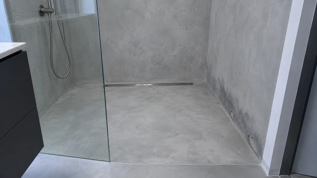 Douche à l'italienne en microciment gris, avec une fissure qui part du caniveau de douche et un pied de mur assombri par l'humidité, avec de petites cloques