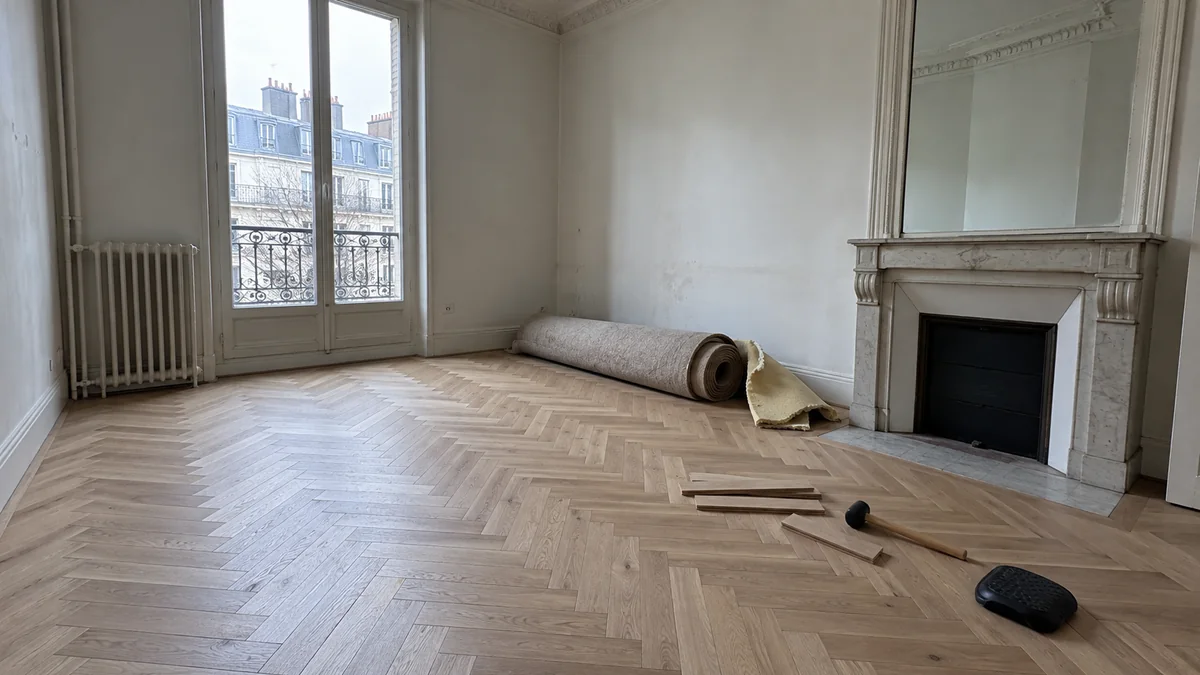 Séjour d'un appartement haussmannien en cours de rénovation : parquet neuf en point de Hongrie posé au sol, ancienne moquette roulée et thibaude jaune déposées contre le mur, maillet de poseur