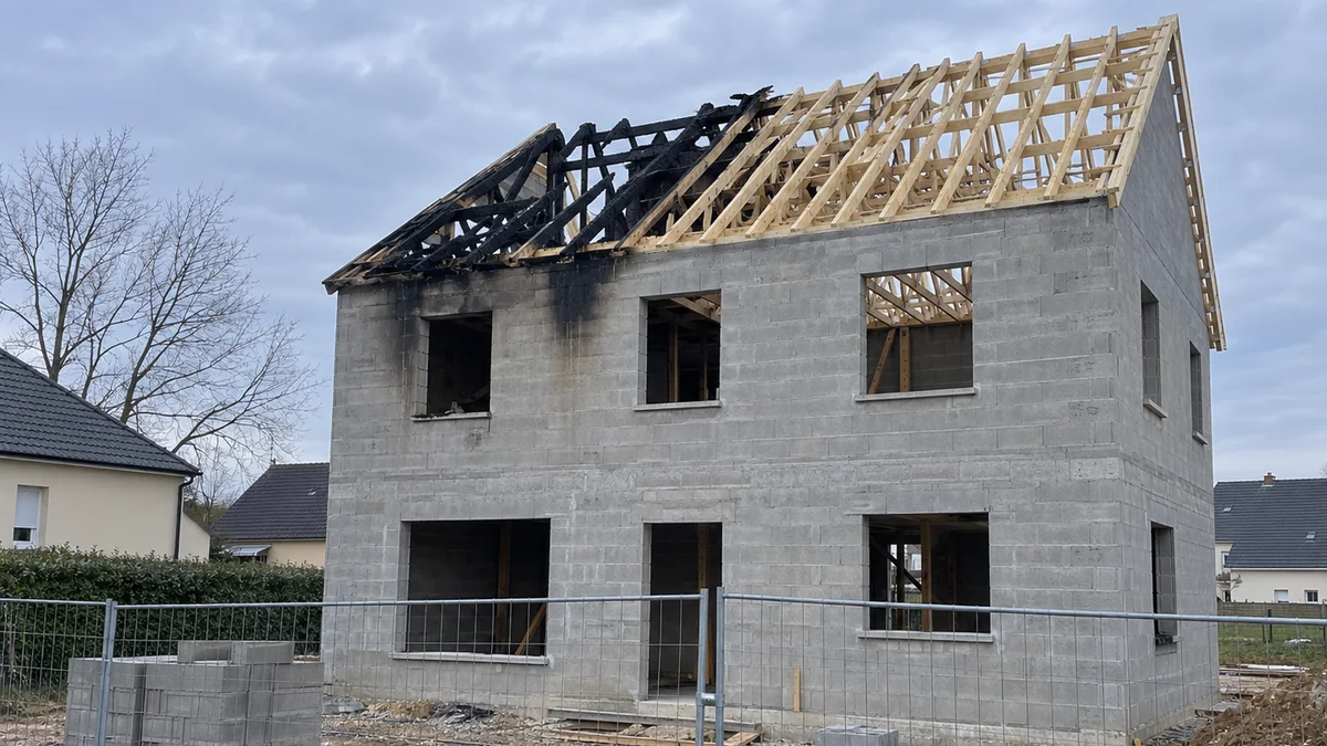 Maison en parpaings en cours de construction, sans menuiseries, dont une partie de la charpente en bois est carbonisée et effondrée après un incendie, traces de suie au-dessus de deux ouvertures, clôture de chantier au premier plan