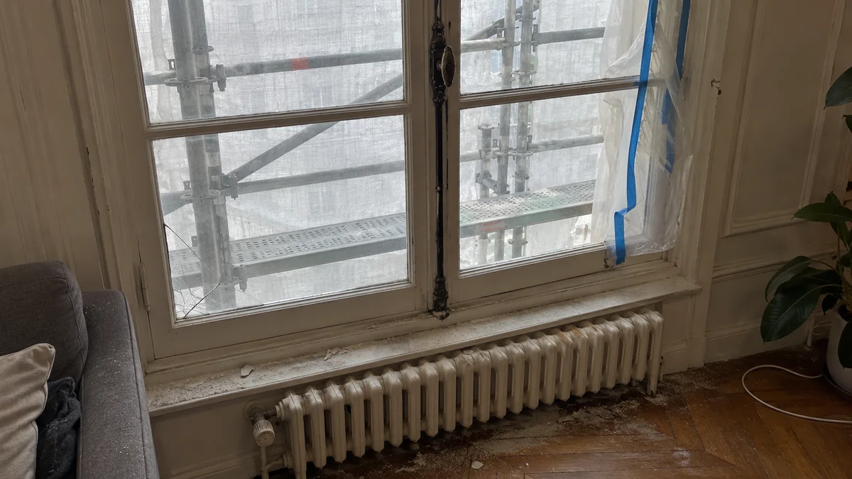 Fenêtre à petits carreaux d'un appartement haussmannien vue de l'intérieur pendant un ravalement : l'échafaudage et son filet gris obscurcissent la vitre, un carreau est fêlé dans l'angle, une fine couche de poussière couvre l'appui et le parquet, du ruban de masquage bleu se décolle du cadre