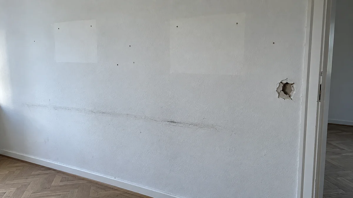 Mur blanc d'un logement vide : deux rectangles plus clairs laissés par des cadres, quelques petits trous de chevilles, une trace grise de meuble et un trou net dans la cloison près de la porte