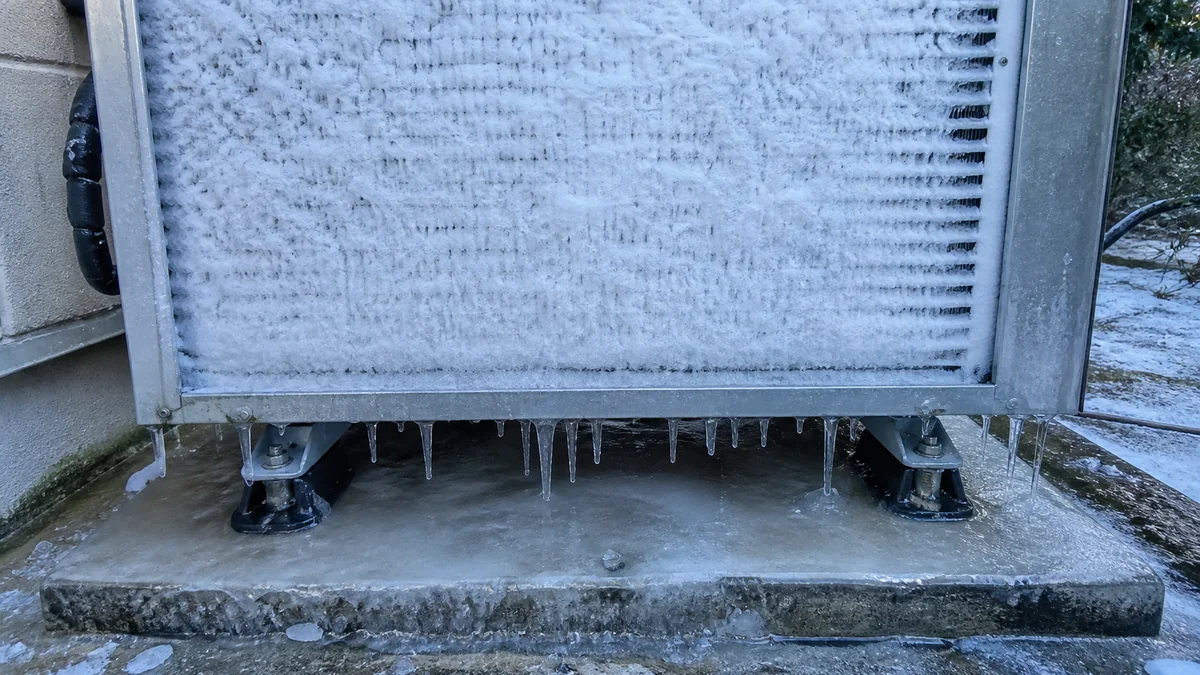 Gros plan sur le bas d'un groupe extérieur de pompe à chaleur : échangeur entièrement pris dans une gangue de givre blanc, stalactites de glace sous le châssis et plaque de glace formée sur le socle béton
