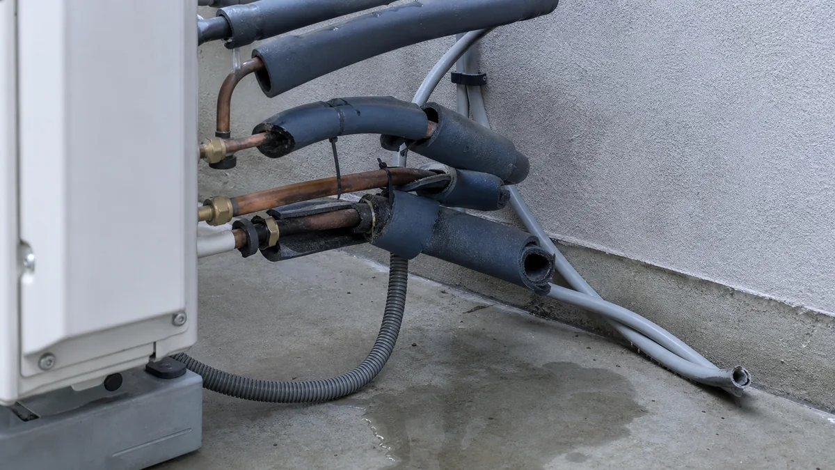 Détail des liaisons frigorifiques en sortie d'un groupe extérieur : gaines isolantes déchirées et béantes laissant les tubes de cuivre à nu, raccords et écrous apparents, tuyau de condensats annelé posé à même le sol