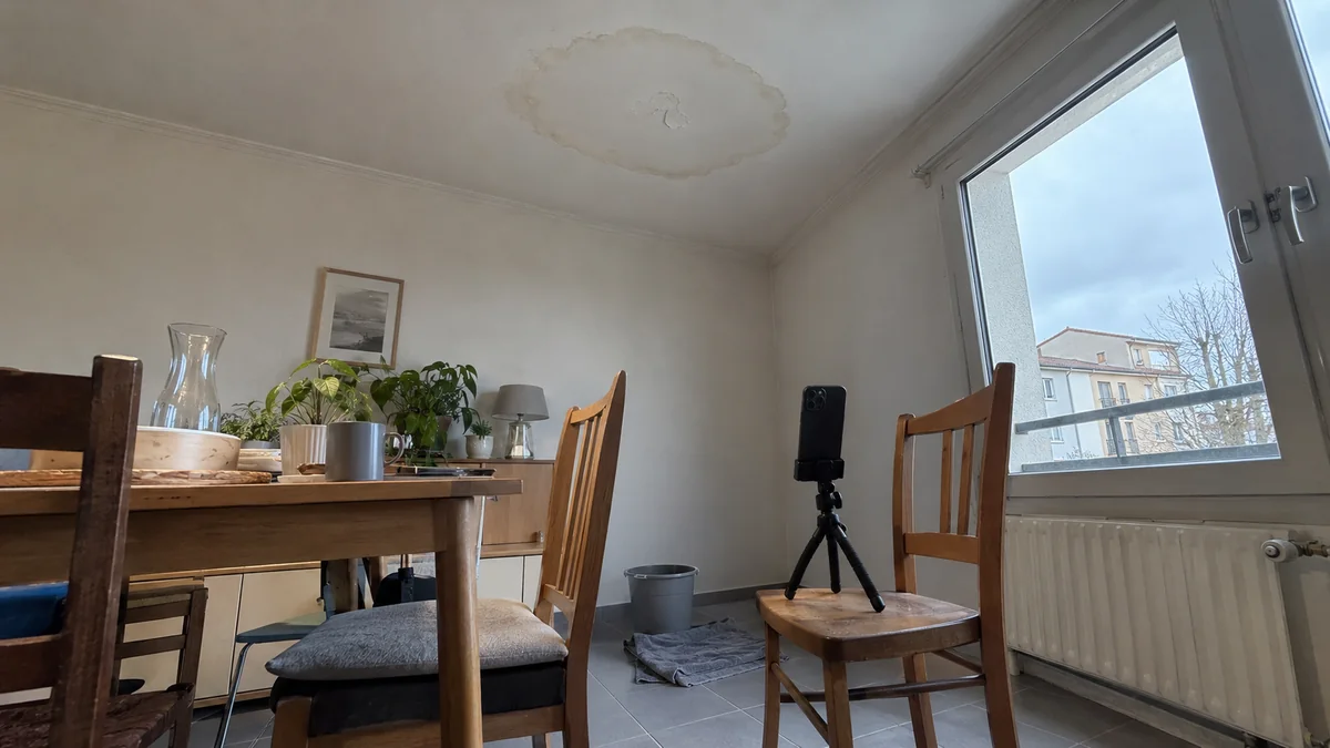 Salle à manger avec une auréole de dégât des eaux au plafond et un smartphone posé sur un trépied pour une expertise en visio, seau et serpillière au sol