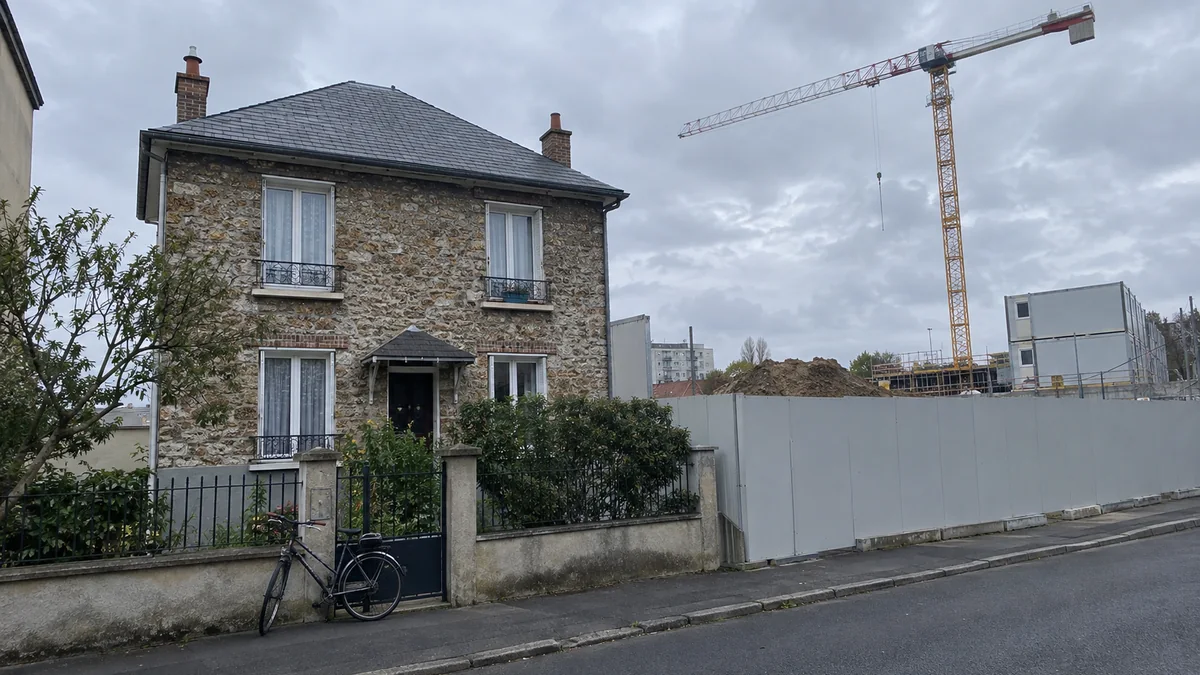 Pavillon en meulière des années 1930 en proche banlieue parisienne, avec juste derrière lui un grand chantier de transport public : grue à tour, palissade grise et bungalows de chantier