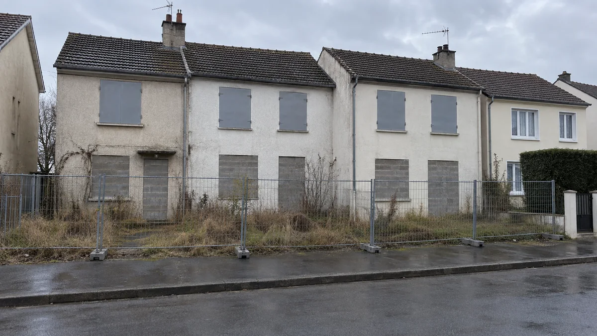 Rangée de petites maisons de banlieue dont les fenêtres et les portes du rez-de-chaussée sont murées en parpaings gris, jardins en friche derrière une clôture de chantier, à côté d'une maison encore habitée