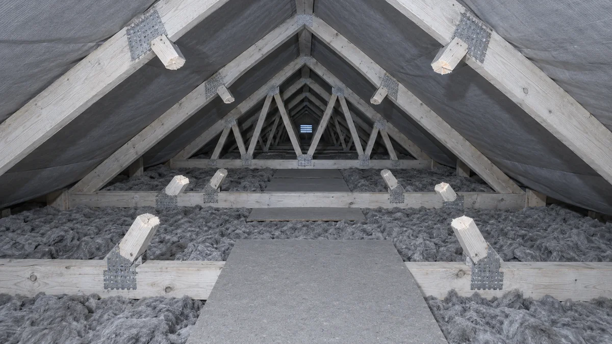 Enfilade de fermettes industrielles dans des combles, dont plusieurs diagonales ont été sciées et ne sont plus que des moignons de bois clair encore fixés aux plaques métalliques crantées, au-dessus d'un isolant en vrac et d'un chemin de panneaux