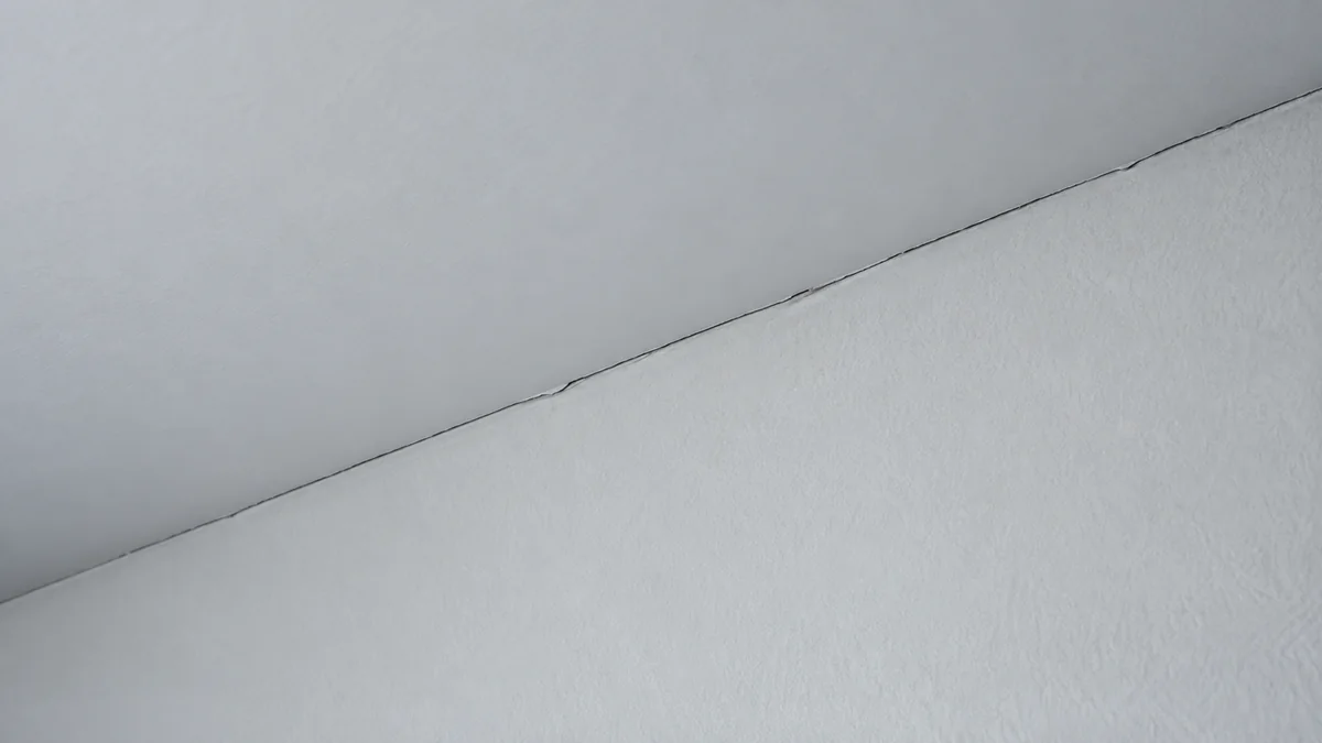 Jonction entre un mur blanc et un plafond blanc dans une chambre, fendue par une fissure fine et continue qui suit exactement l'angle