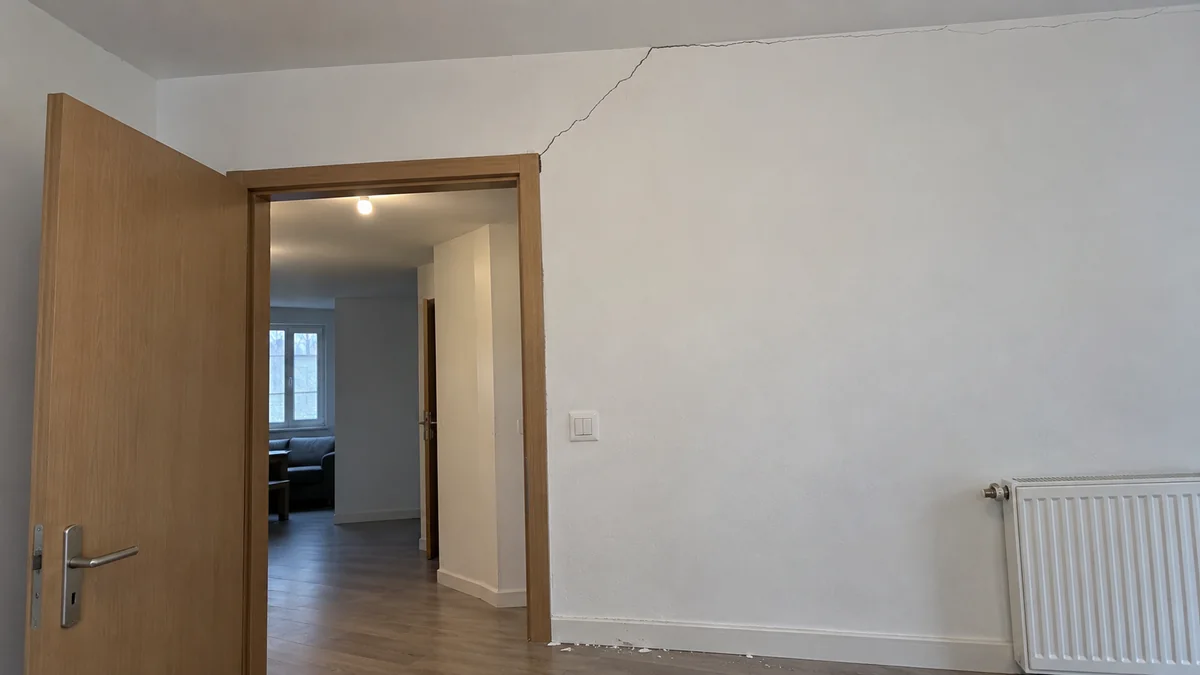 Mur blanc d'une pièce autour d'une porte intérieure en bois ouverte sur le couloir : une fissure fine part de l'angle supérieur de l'huisserie et monte en diagonale vers le plafond, quelques écailles de peinture sont tombées sur la plinthe, une fine fissure court aussi le long de la jonction entre le mur et le plafond
