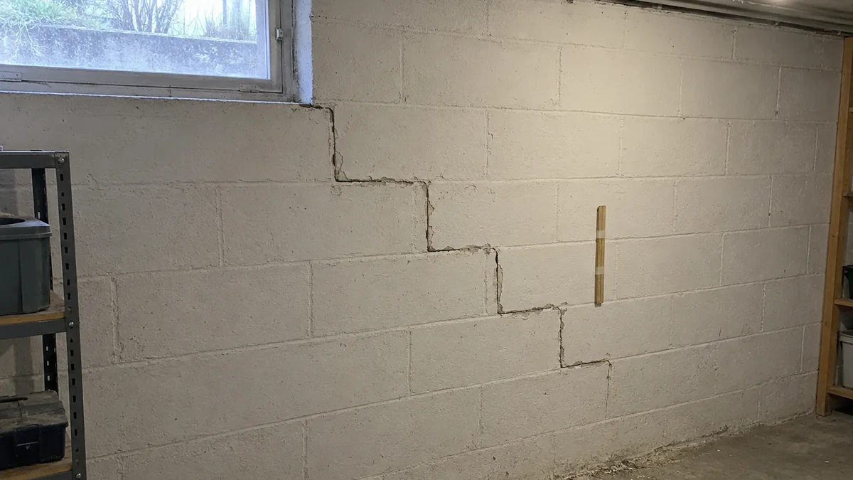 Mur de cave en blocs de béton peints en blanc : une fissure en escalier suit les joints de mortier depuis le soupirail sur près d'un mètre et demi, un mètre pliant en bois est scotché à côté pour l'échelle, sol en béton