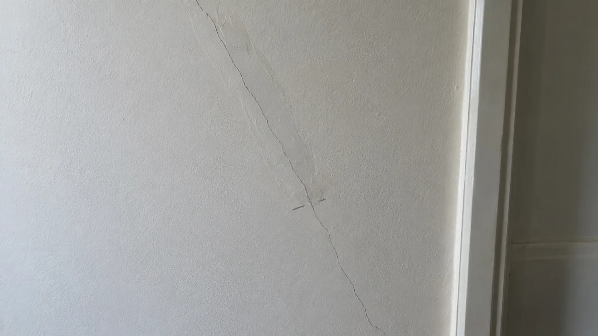 Mur intérieur blanc près d'un encadrement de porte : une fissure en biais traverse une ancienne bande de rebouchage encore visible en relief, puis se prolonge au-delà de cette bande par une fissure fine et nette ; deux petits traits de crayon marquent la fin de l'ancienne réparation