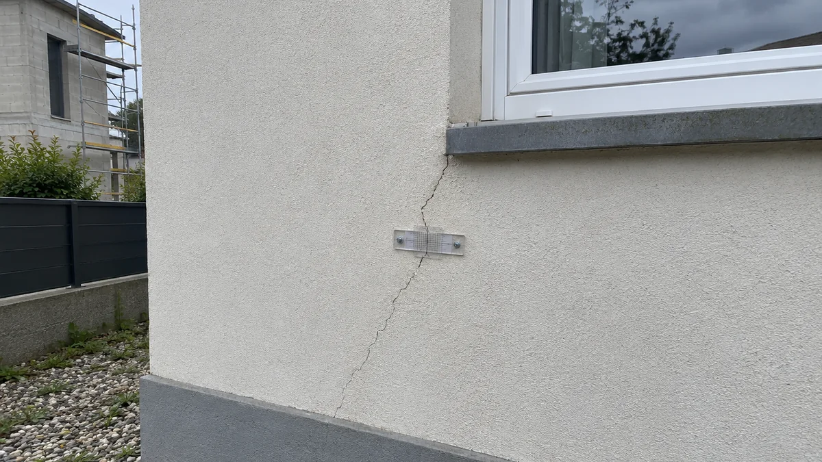 Façade enduite d'une maison individuelle : une fissure part de l'angle bas d'une fenêtre et descend en biais jusqu'au soubassement gris, un témoin de fissure en plastique transparent est vissé en travers ; à gauche, derrière la clôture, un bâtiment voisin en construction avec son échafaudage