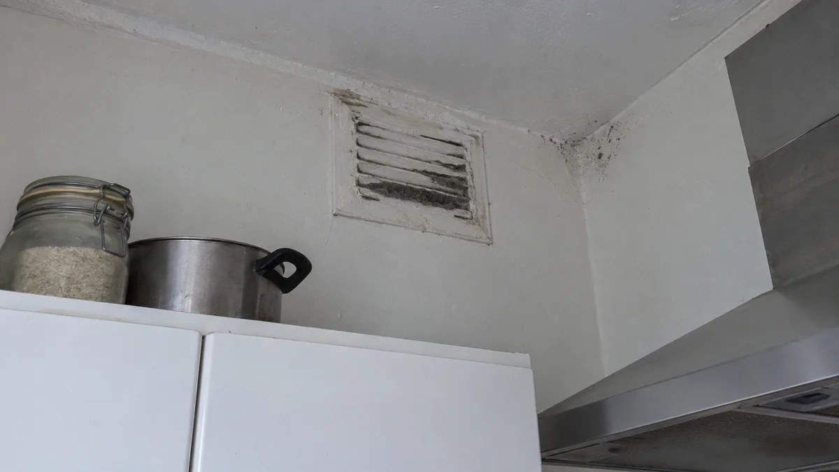Grille de ventilation haute d'une cuisine ancienne repeinte plusieurs fois et encrassée de poussière grasse, avec des points de moisissure dans l'angle du plafond au-dessus des placards