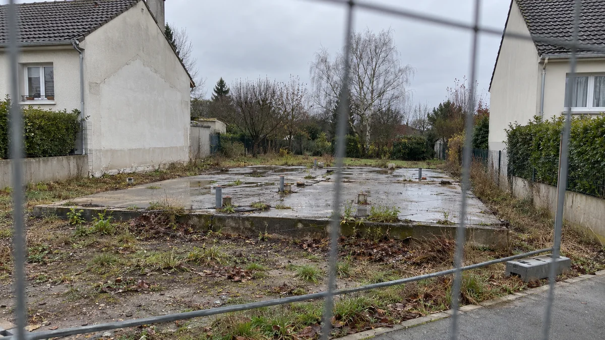Terrain vide entre deux maisons intactes après la démolition d'une maison sinistrée : dalle de béton nue avec des tuyaux d'évacuation coupés, flaques et herbes, clôture de chantier au premier plan