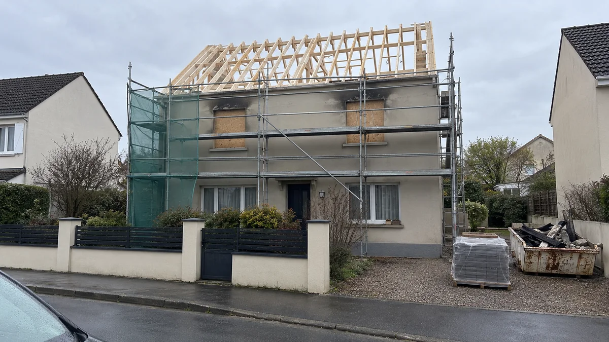 Maison en reconstruction après un incendie : charpente neuve en bois clair posée sur les murs d'origine, traces de suie au-dessus des fenêtres condamnées, échafaudage et palette de tuiles dans l'allée