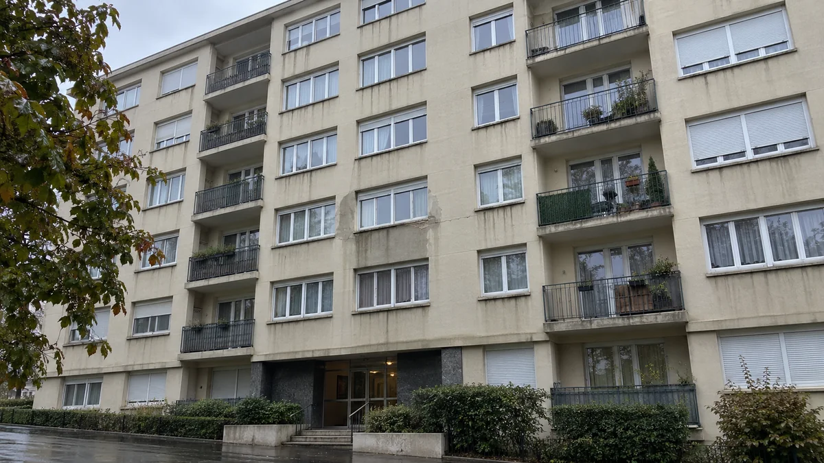 Façade d'un immeuble de logements sous la pluie, enduit beige marqué de coulures sombres et d'une longue fissure verticale entre deux rangées de fenêtres