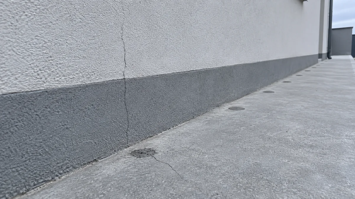 Pied de façade d'une maison avec une rangée de trous d'injection rebouchés au mortier dans le trottoir en béton, une fissure fine partant d'un bouchon et une autre remontant dans l'enduit au-dessus du soubassement