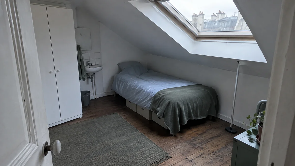 Chambre de bonne sous les toits à Paris vue depuis la porte : plafond en forte pente vers une fenêtre de toit, lit simple glissé sous la partie basse, petit lavabo et armoire blanche