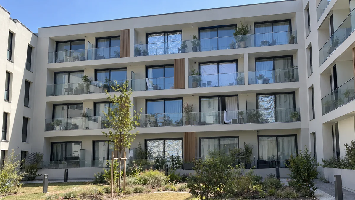 Façade d'une résidence neuve exposée au soleil d'après-midi : grandes baies sans volets ni stores extérieurs, draps et films réfléchissants posés par les habitants derrière les vitres, une gaine de climatiseur mobile sortant d'une fenêtre entrouverte