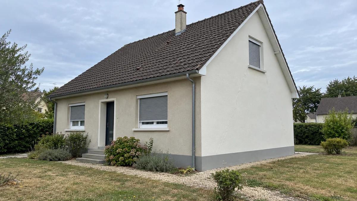 Pavillon des années 1960 en grande banlieue vu depuis le jardin : la façade avant à l'enduit beige un peu terne contraste avec le pignon fraîchement repeint en blanc cassé, plus clair, et sous la peinture neuve du pignon se devinent de longues lignes de fissures rebouchées qui descendent en biais depuis la fenêtre des combles vers le soubassement gris