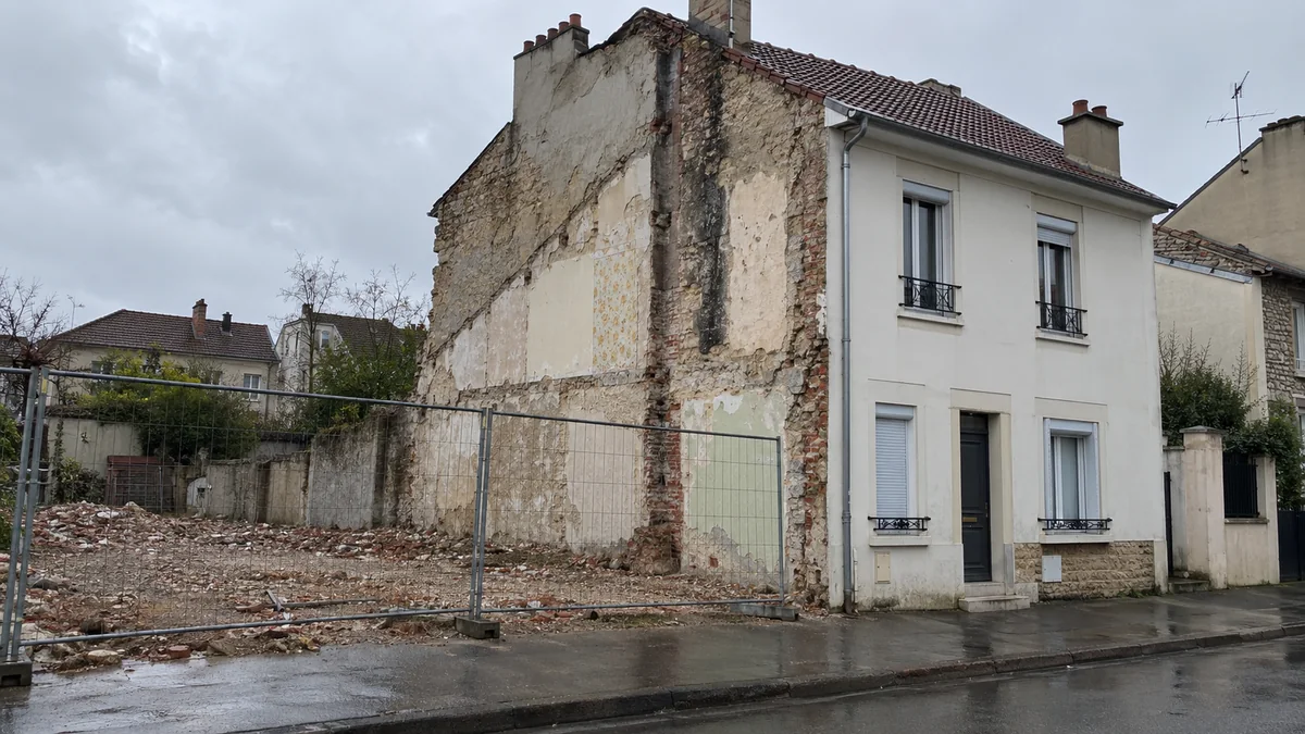 Maison de ville à deux niveaux vue depuis le trottoir après la démolition de la maison accolée : le mur de côté désormais à nu montre des pans de plâtre et de papier peint délavé, la brique et la meulière apparentes, une bande sombre de suie et d'humidité, devant un terrain de gravats fermé par des barrières de chantier grillagées