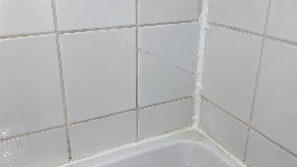 Faïence murale autour d'une baignoire : un carreau fendu et un joint silicone étalé grossièrement dans l'angle