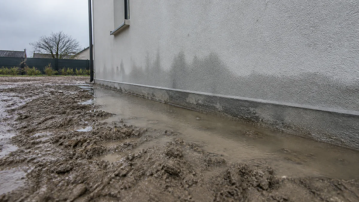 Eau de pluie stagnant contre le pied d'un mur de fondation en béton après une averse, signe d'un défaut de pente ou de drainage
