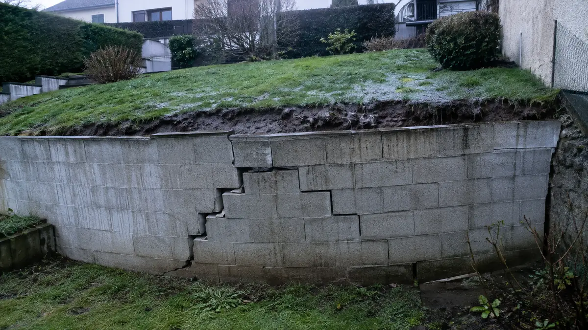 Mur de soutènement en parpaings penché vers l'avant, ventre marqué et fissure en escalier dans la maçonnerie