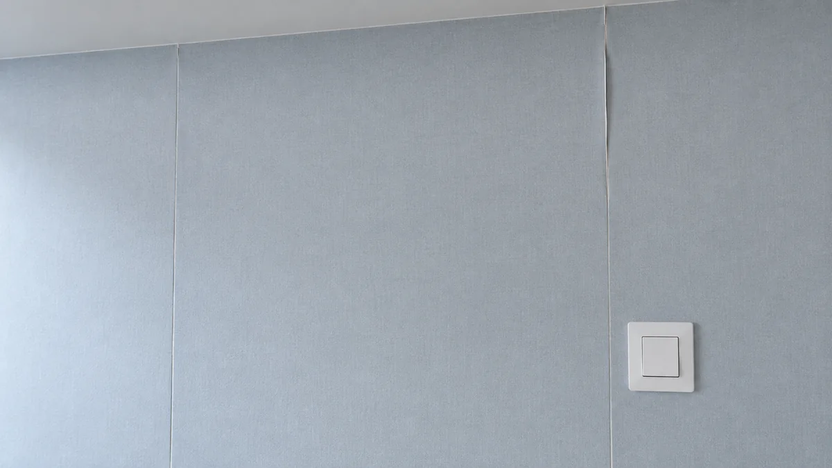 Mur tapissé d'un papier peint gris bleu dont deux joints verticaux se sont ouverts de quelques millimètres, l'un se relevant près du plafond