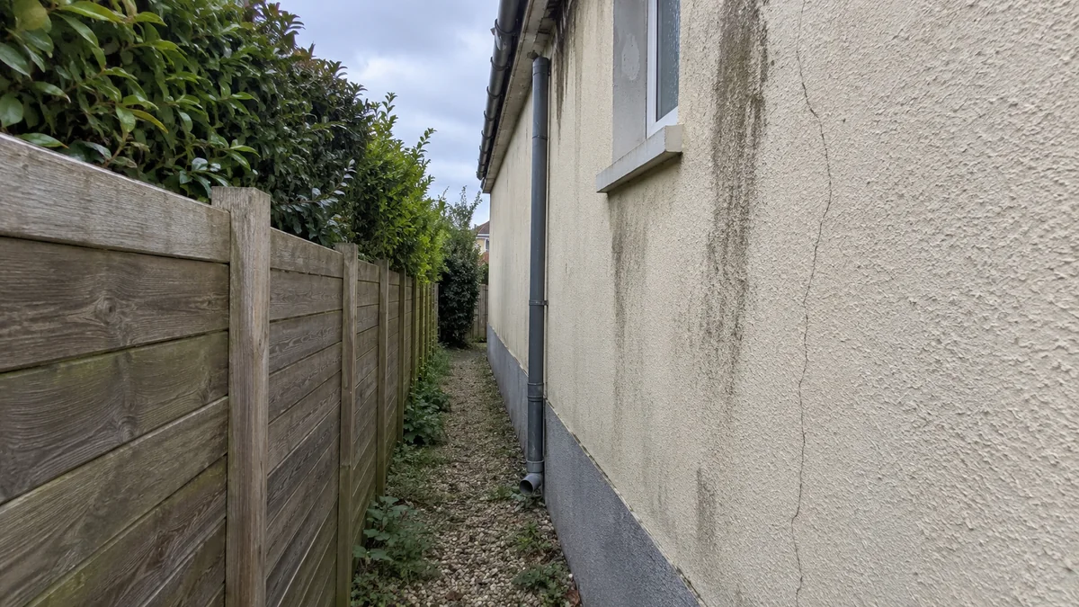 Étroit passage d'une soixantaine de centimètres entre le mur latéral d'une maison et la clôture en bois du voisin, sol gravillonné envahi d'herbes : l'enduit crème du mur est strié de coulures sombres sous la gouttière et parcouru d'une longue fissure verticale, trop près de la clôture pour y monter un échafaudage