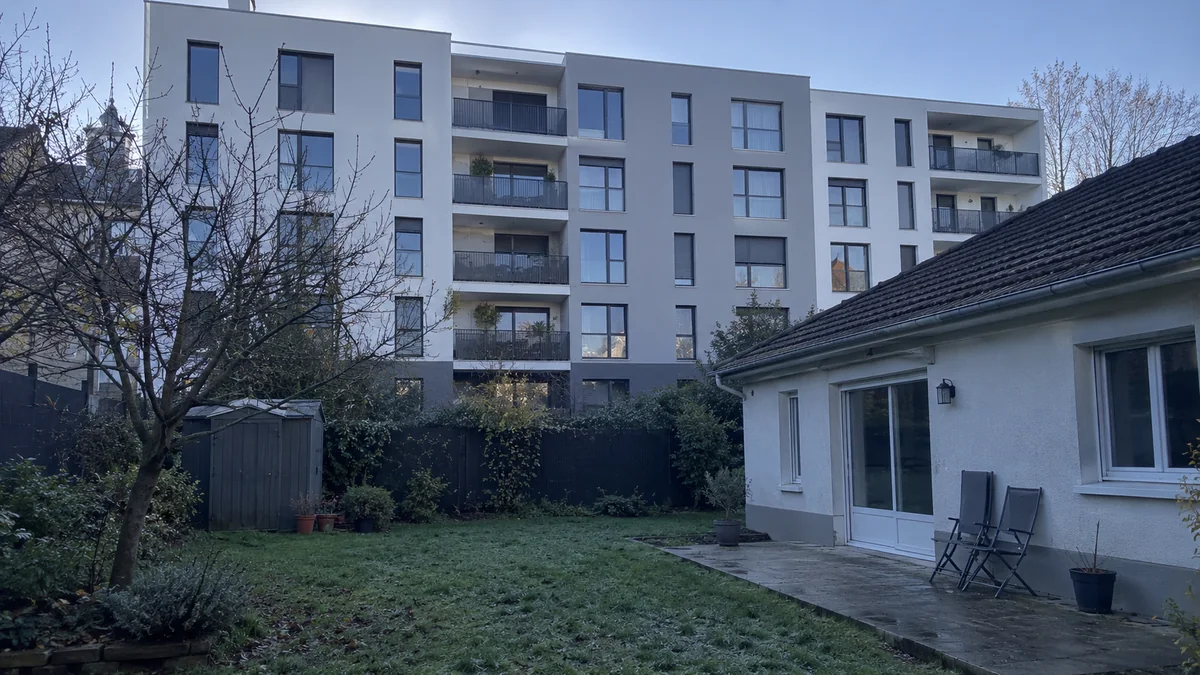 Jardin d'un pavillon de banlieue parisienne en hiver, pelouse givrée et terrasse avec deux chaises pliantes, dominé à quelques mètres de la clôture du fond par un immeuble neuf de six niveaux aux balcons métalliques qui plonge le jardin et la façade de la maison dans l'ombre