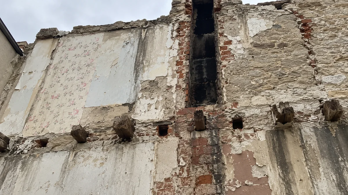 Face extérieure d'un ancien mur mitoyen vue d'en bas après la démolition de la maison voisine : bande de papier peint fleuri délavé, conduit de cheminée en brique coupé et noirci de suie, rangée d'abouts de solives en bois encore pris dans la maçonnerie et plâtre qui s'effrite sur la brique et la meulière