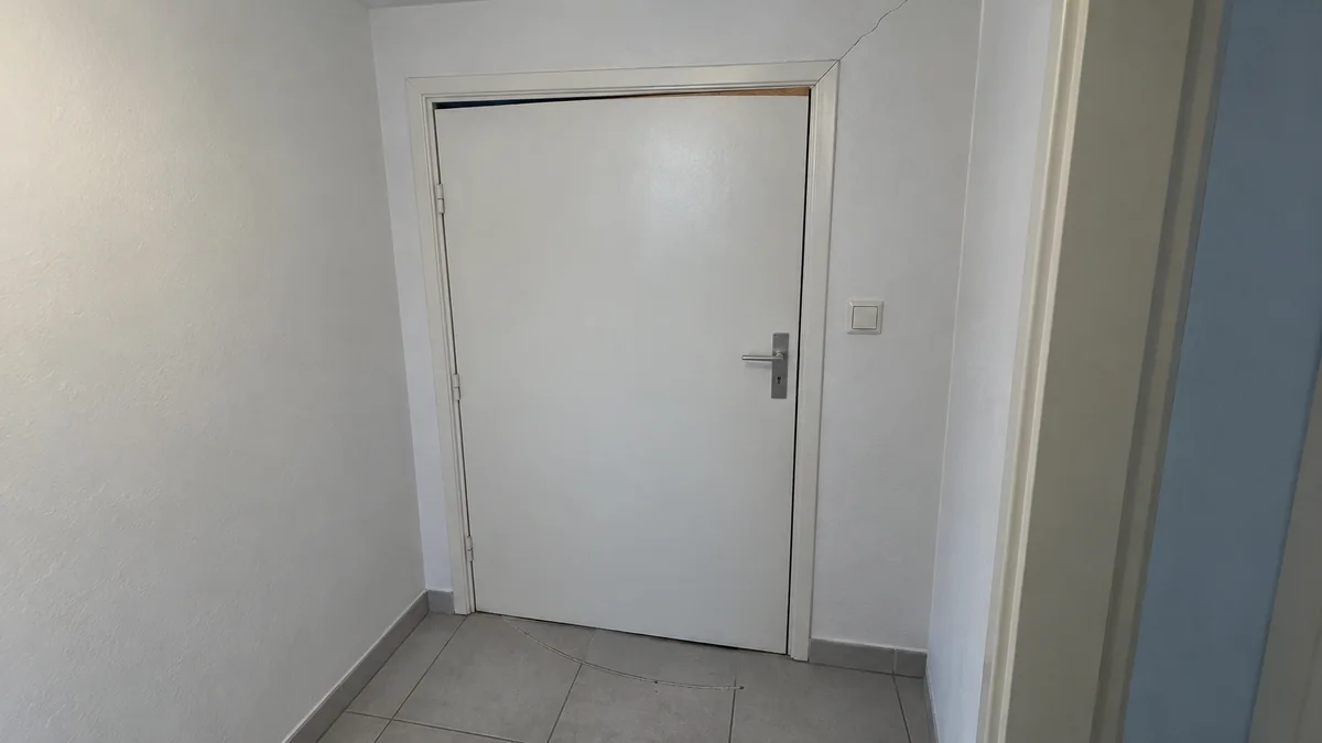 Porte intérieure blanche dans un couloir carrelé : le jeu entre le haut de la porte et l'huisserie s'ouvre légèrement en biais, une fine bande de bois brut apparaît sur le chant supérieur raboté, une fine fissure part de l'angle supérieur de l'huisserie vers le plafond, et une trace en arc marque le carrelage là où la porte frotte