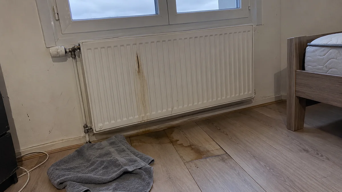Radiateur à panneau en acier blanc sous une fenêtre de chambre, fendu sur un des canaux verticaux avec une traînée brunâtre sur la peinture, une tache séchée sur le parquet stratifié en dessous et une serviette grise pliée au sol