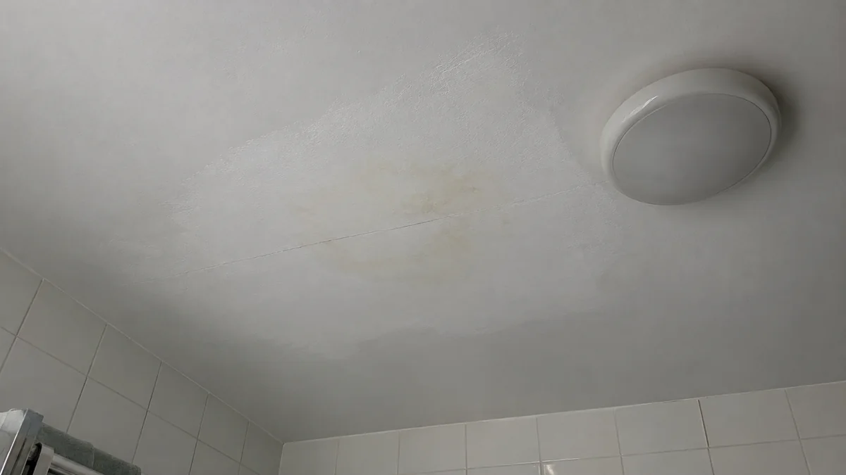Plafond de salle de bains vu d'en bas après une réparation organisée par l'assurance : la zone repeinte se distingue par sa texture de rouleau et son contour irrégulier, une auréole jaunâtre réapparaît au centre à travers la peinture neuve, traversée par une fine fissure le long d'un joint de plaque, à côté d'un plafonnier rond