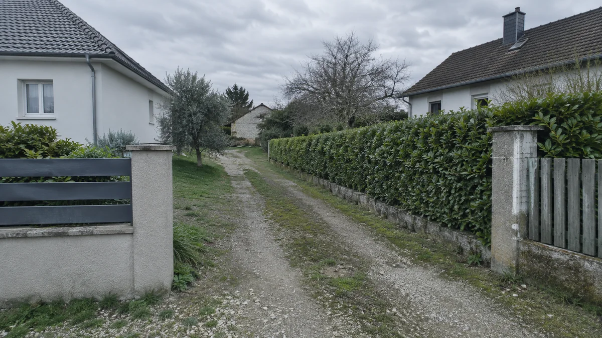 Chemin de terre traversant en diagonale deux propriétés voisines délimitées par une haie basse, vue depuis l'entrée du terrain