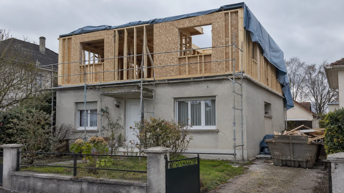 Pavillon de plain-pied crépi gris en cours de surélévation : un étage en ossature bois et panneaux OSB est monté sur les murs existants et couvert en partie par une bâche bleue, un échafaudage longe la façade et une benne remplie de chutes de bois occupe l'allée