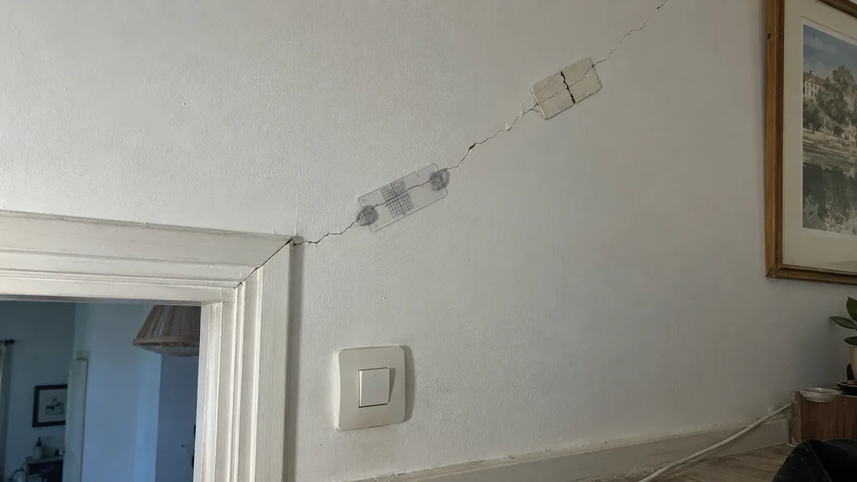Mur intérieur blanc fendu en diagonale depuis l'angle d'un encadrement de porte : une jauge de fissure en plastique transparent à quadrillage est collée en travers de la fissure, et plus haut un témoin en plâtre rectangulaire s'est rompu en son milieu