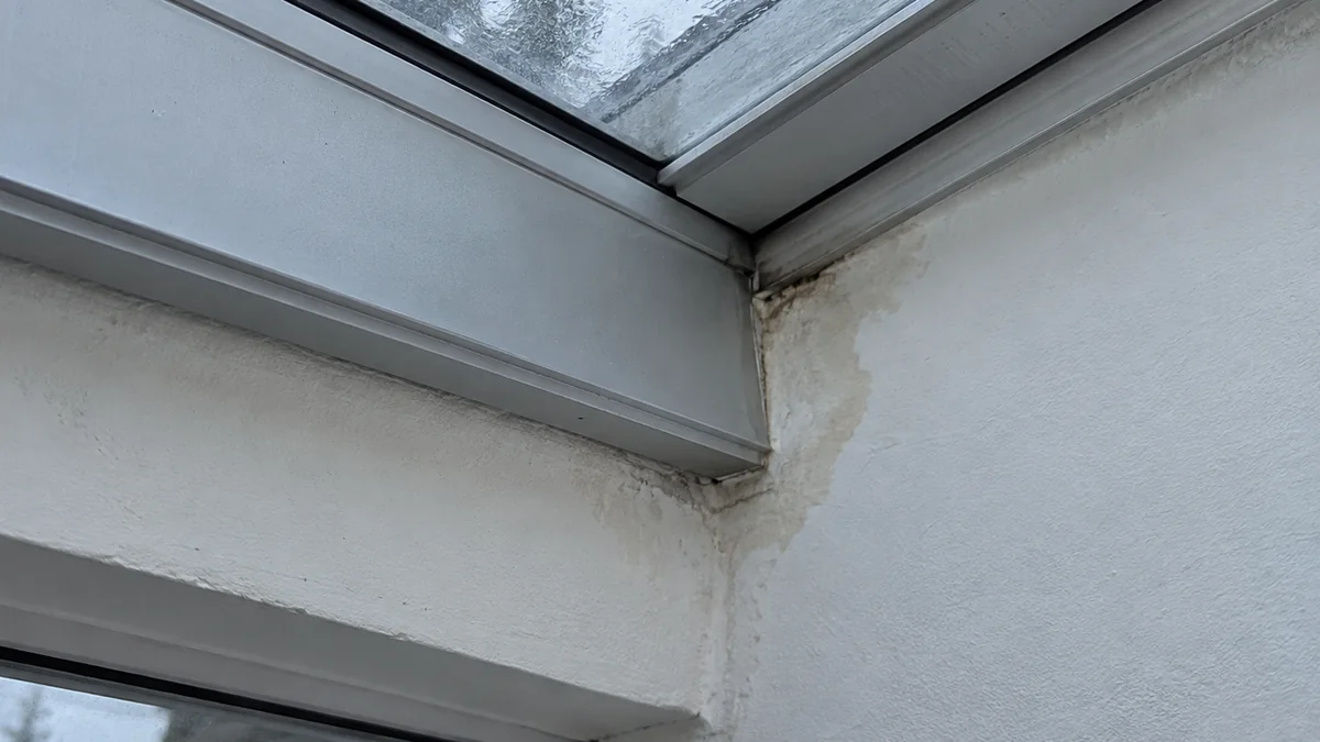 Tache d'infiltration brunâtre visible au plafond, à la jonction entre la toiture vitrée d'une véranda et le mur de la maison