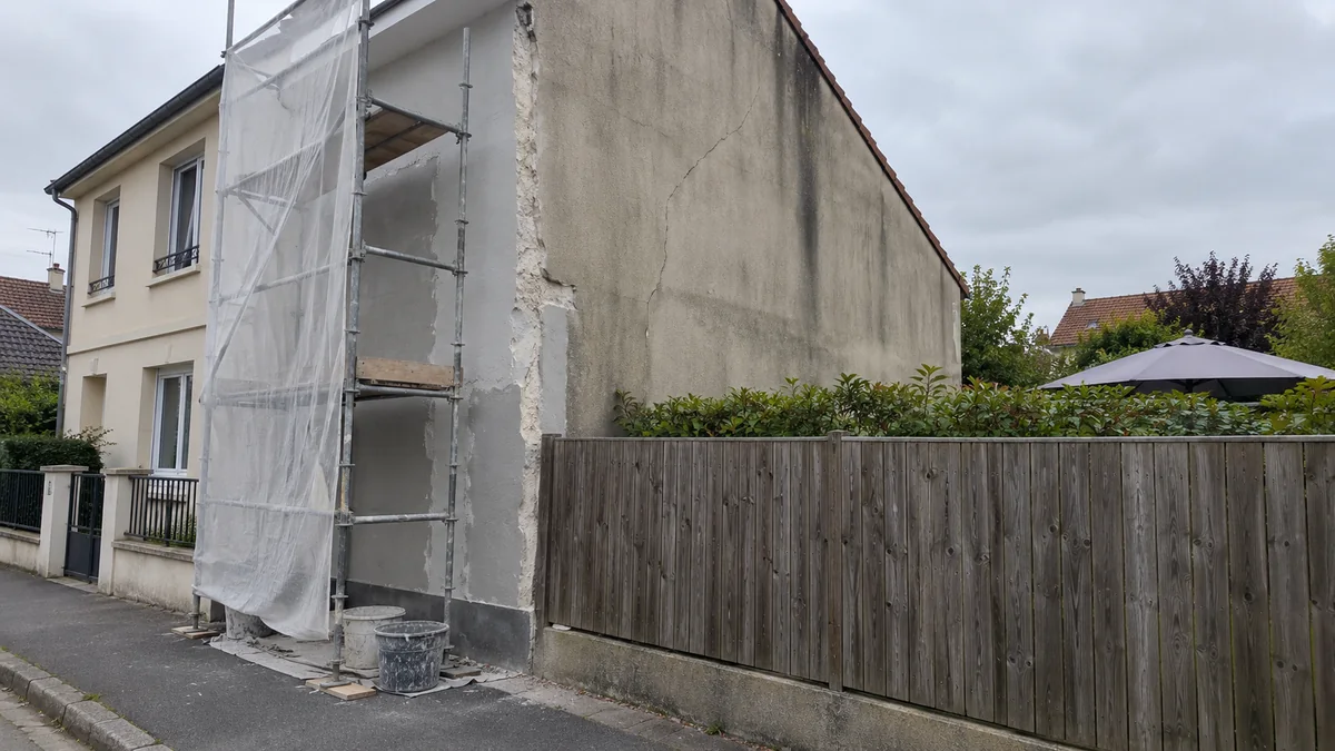 Pignon d'une maison ancienne bâti en limite de propriété : un échafaudage habillé d'un filet blanc couvre seulement la partie côté rue et s'arrête net à la clôture en bois du voisin, le reste du mur à l'enduit taché et fissuré restant hors de portée derrière la haie du jardin voisin