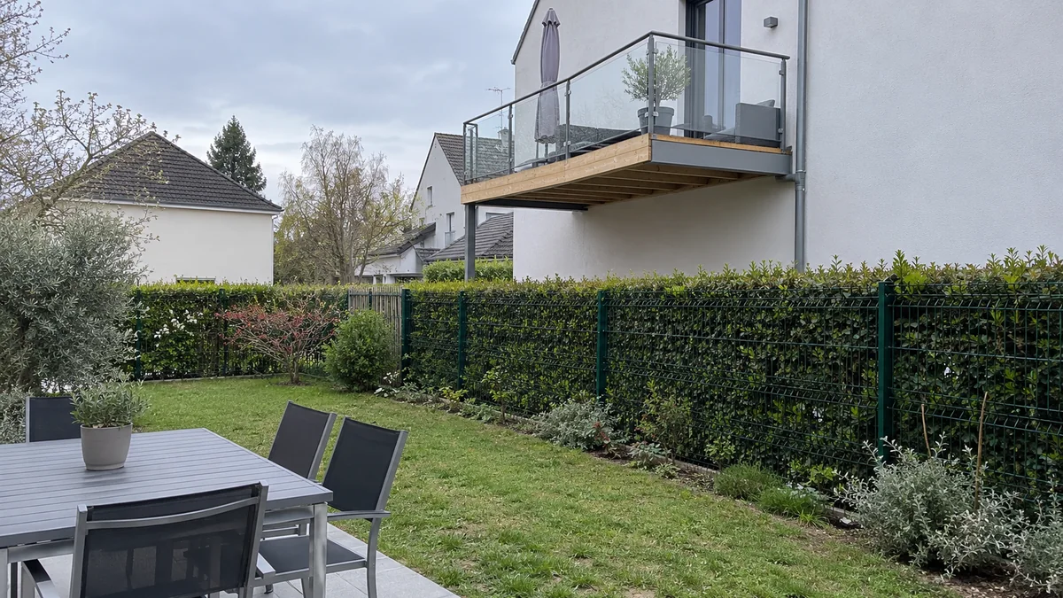 Jardin d'une maison avec table et chaises, bordé d'une clôture grillagée doublée d'une haie ; juste derrière, le balcon neuf du voisin en bois et garde-corps vitré surplombe la limite et plonge sur la pelouse