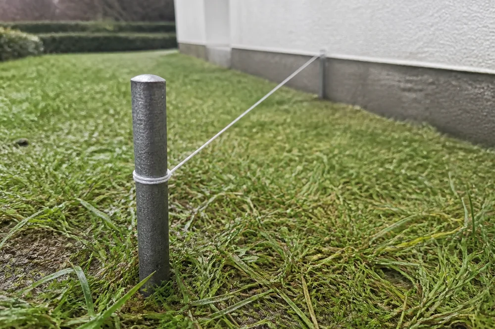 Borne de délimitation métallique plantée dans une pelouse, avec un cordeau tendu vers la fondation d'une maison en construction, aucune personne visible